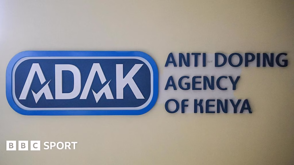 The blue and white logo of the Anti-Doping Agency of Kenya, ADAK, is displayed on a wall with the full text of the organisation's name to the right of the logo