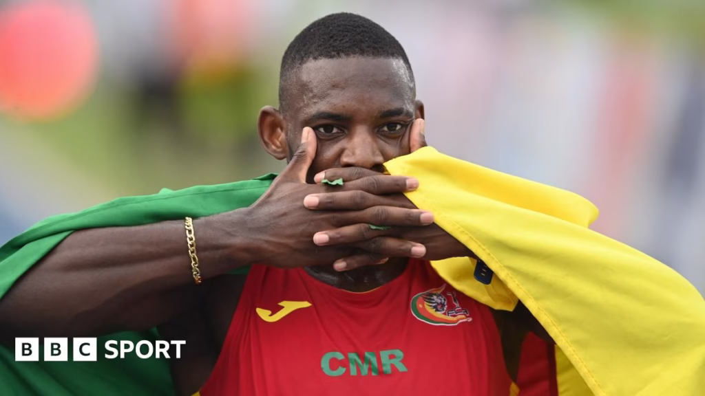 Emmanuel Eseme, seen from chest up wearing a vest with green and white horizontal stripes and a Sporting Clube de Portugal crest on it, covers his mouth with interlocking fingers to mimic the goal celebration of Arsenal striker Viktor Gyokeres. A row of bright lights and an athletics track are seen blurred in the background
